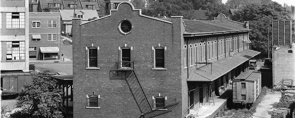 A brick warehouse with an iron fire escape beside a rail siding, boxcars at the loading dock.