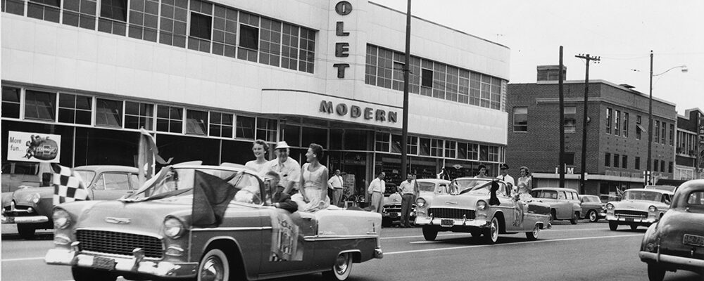 A parade of new 1955 Chevrolet convertibles with riders waving, passing the Modern Chevrolet showroom.