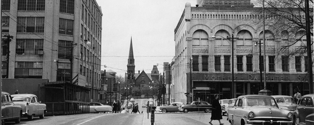 A downtown Winston-Salem street in the 1950s, cars parked along both curbs and a church steeple at the end of the block.